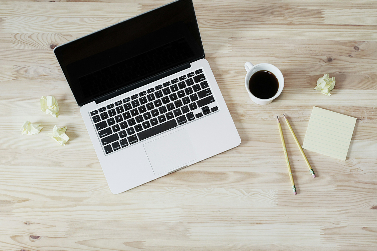 Overhead view of desk with laptop, coffee, and notepad.